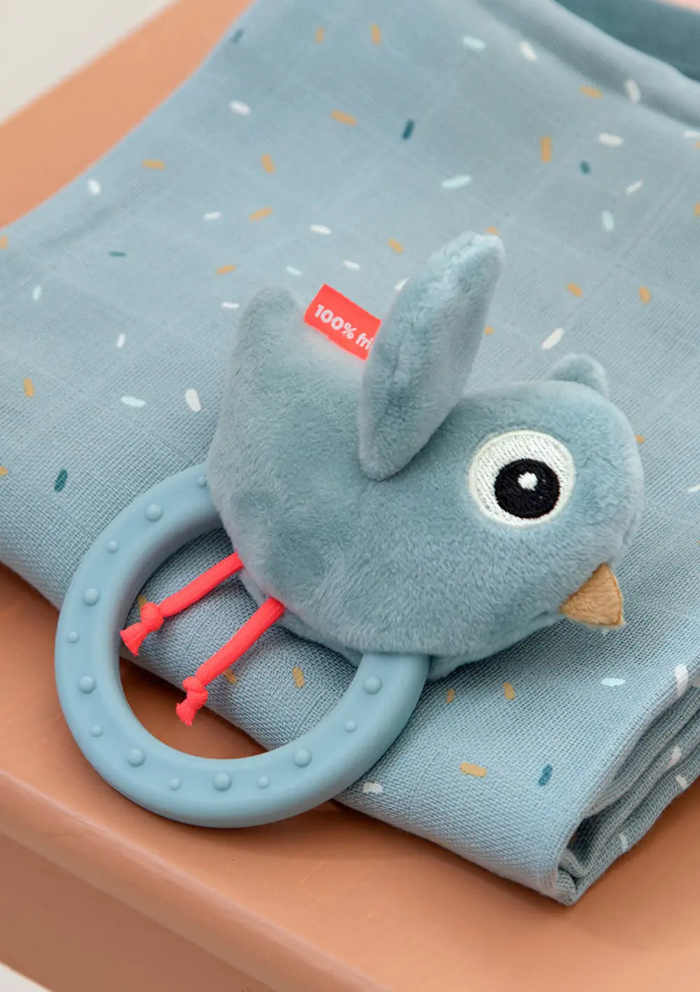 Done by Deer Rassel mit Beißring 'Birdee' blau liegt auf einem gemustertem Mulltuch.