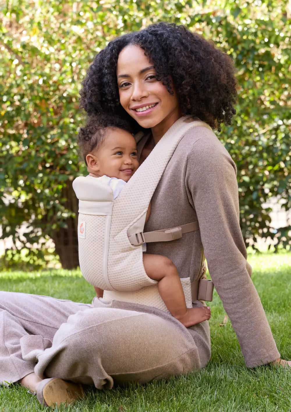 Frau sitzt auf einer Wiese und trägt ihr Baby in der Ergobaby Babytrage 'Omni Lite' Mesh Natural Beige