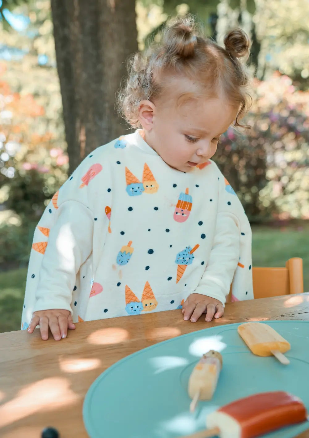 Nahaufnahme eines Kindes, das im cremeweißen Little Casimir Langarm-Lätzchen Boo Ice Cream am Holztisch steht und neugierig auf verschiedene Sorten Eis am Stiel blickt.