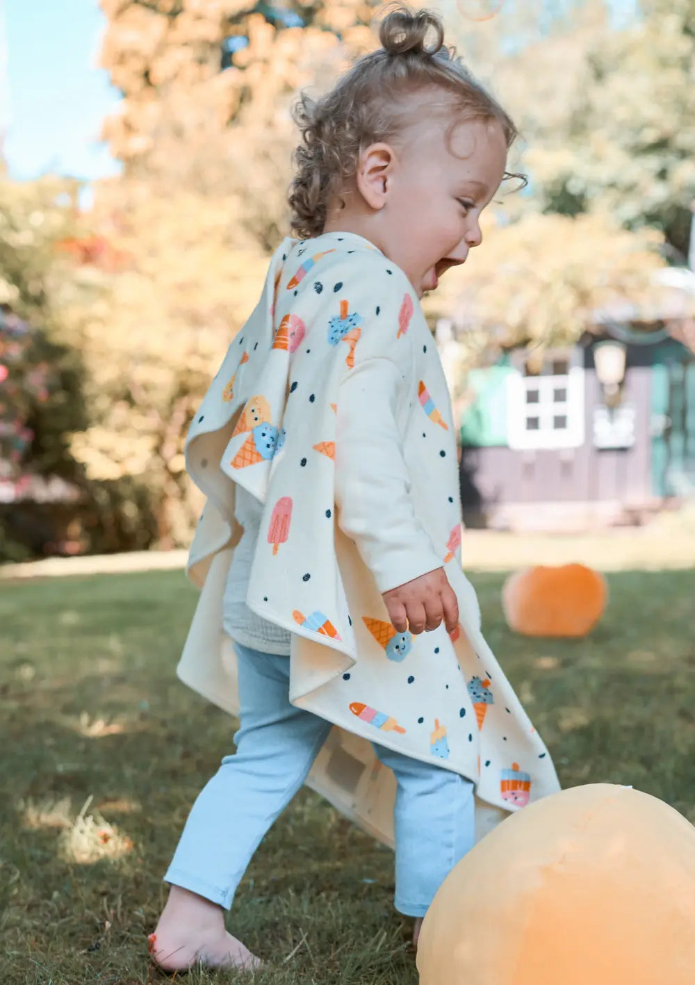 Ein fröhliches Kleinkind läuft barfuß im Garten über den Rasen und trägt das cremeweiße Little Casimir Langarm-Lätzchen Boo mit buntem Eiswaffel-Print über der Kleidung.
