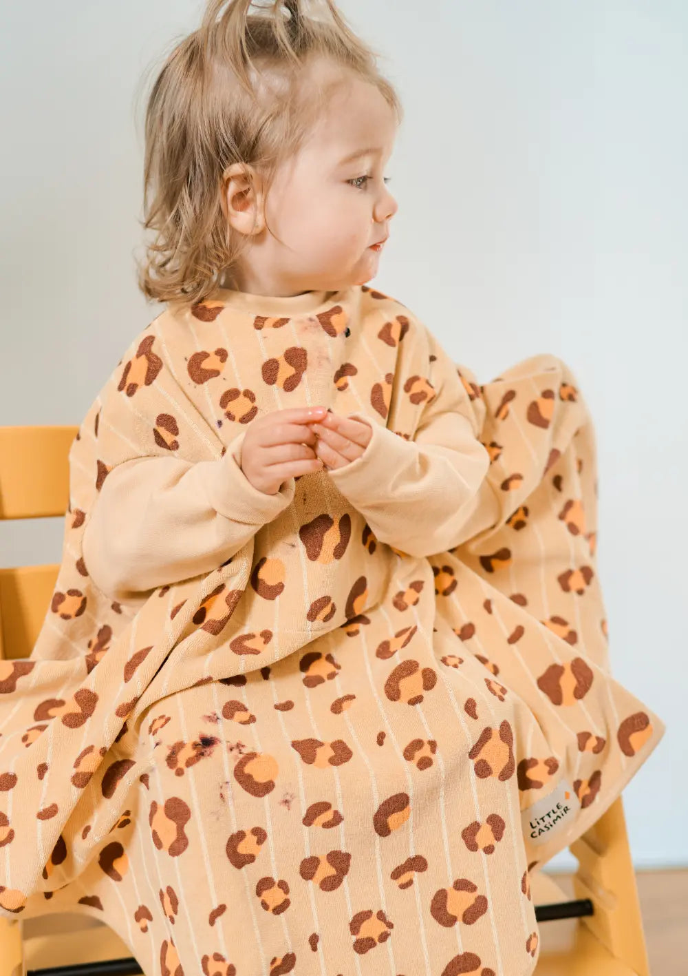 Ein kleines Mädchen sitzt im Hochstuhl und trägt das honiggelbe Little Casimir Langarm-Lätzchen Boo Honey Haze, das mit feinen Längsstreifen und einem braun-orangefarbenen Leoparden-Muster versehen ist.