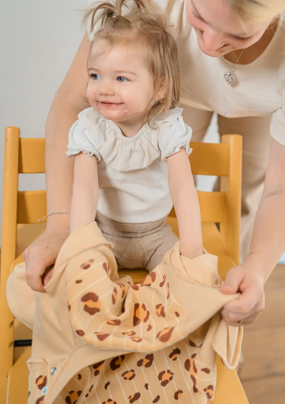 Eine Mutter zieht ihrer lächelnden Tochter im Hochstuhl das verschmutzte, honiggelbe Little Casimir Langarm-Lätzchen Boo Honey Haze nach dem Essen vorsichtig über die Arme aus.