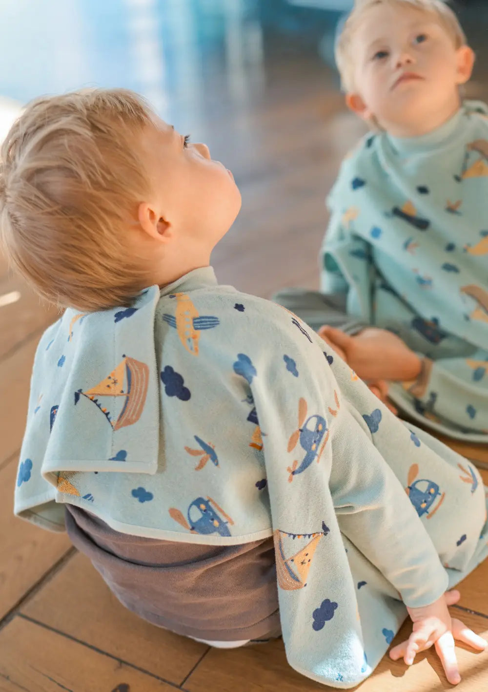 Ein Kind sitzt auf dem Holzboden vor einem großen Spiegel und trägt das hellblaue Little Casimir Langarm-Lätzchen Boo Little Voyagers, das rundherum mit kleinen Fahrzeugmotiven wie Schiffen und Flugzeugen gestaltet ist.