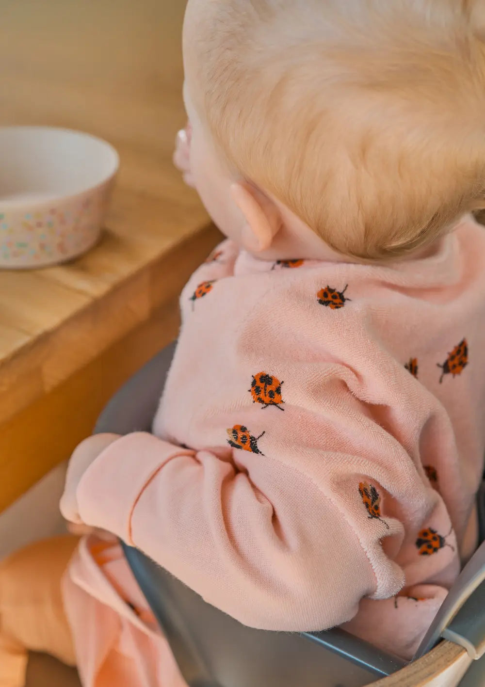 Ein kleines Kind sitzt am Esstisch und greift fröhlich nach seiner Schale, optimal geschützt durch das weiche, rosa Little Casimir Langarm-Lätzchen Boo Love Bugs mit Marienkäfer-Print.