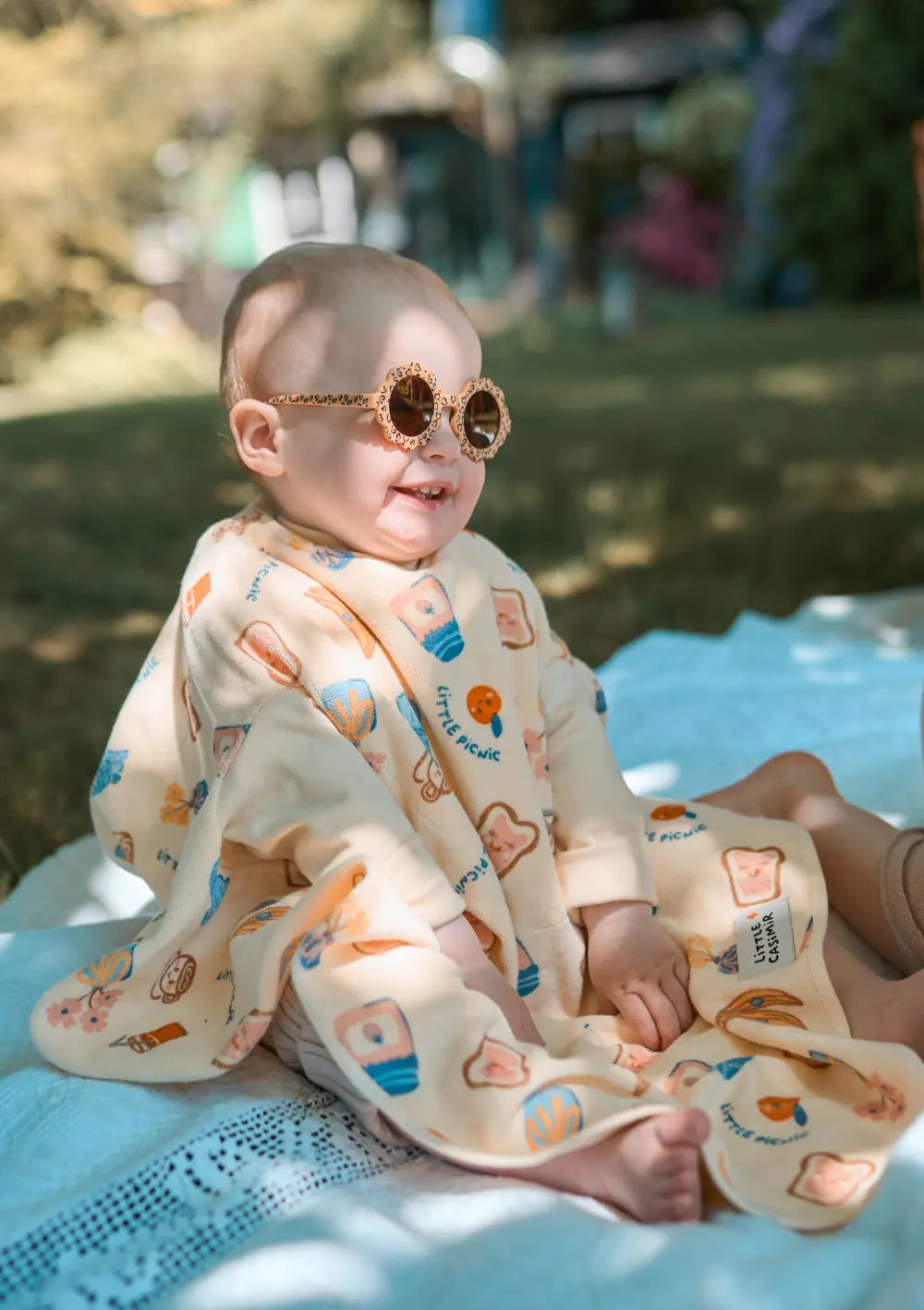 Ein lächelndes Kleinkind sitzt mit einer gemusterten Sonnenbrille im Garten auf einer Picknickdecke und trägt das hellgelbe Little Casimir Langarm-Lätzchen Boo Picnic Club mit verspielten Picknick-Motiven wie Toastscheiben und Früchten.