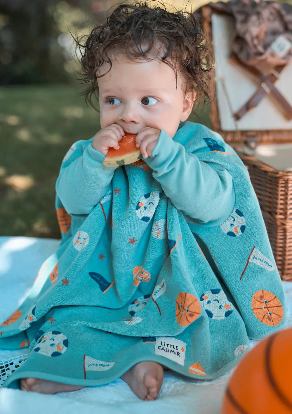Ein Kind mit lockigem Haar sitzt im Freien auf einer Picknickdecke und isst ein Stück Wassermelone, optimal geschützt durch das türkisfarbene Little Casimir Langarm-Lätzchen Boo Tiny Champs mit buntem Sport-Print.