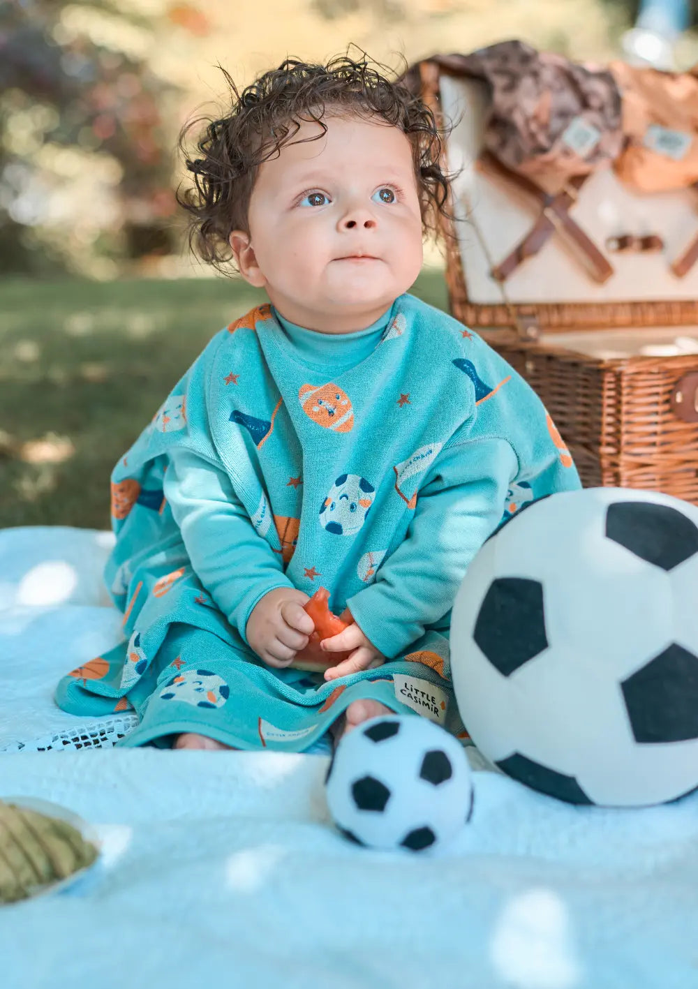 Ein Kleinkind sitzt bei einem Picknick auf einer Decke im Gras und trägt das türkisfarbene Little Casimir Langarm-Lätzchen Boo Tiny Champs mit sportlichen Ballmotiven, während zwei Plüsch-Fußbälle vor ihm liegen.