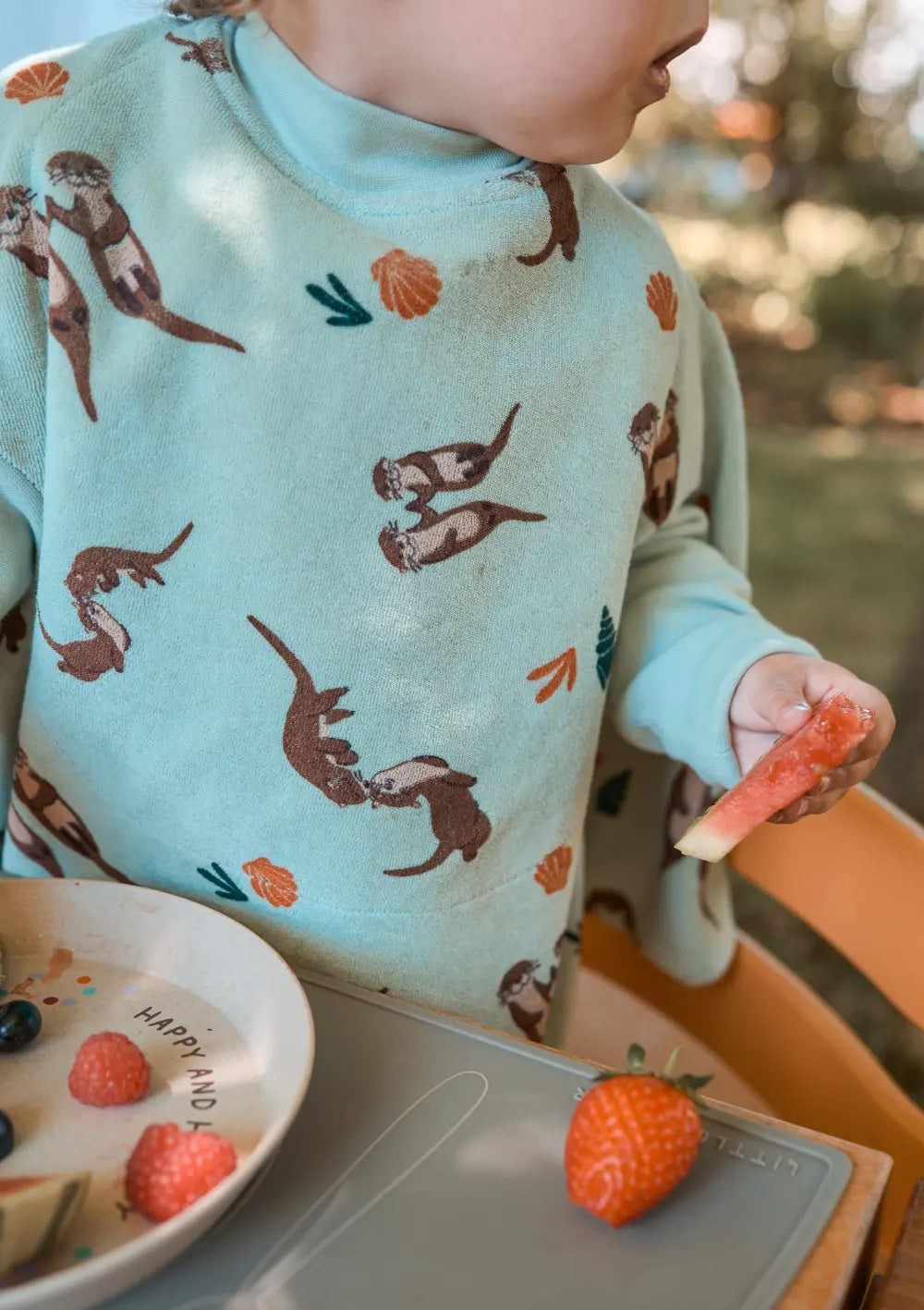 Nahaufnahme eines Kindes, das im Little Casimir Langarm-Lätzchen Boo Otter am Tisch sitzt und ein saftiges Stück Wassermelone in der Hand hält, während auf der Platzmatte eine frische Erdbeere liegt.