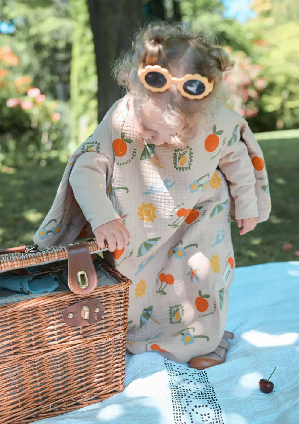 Ein Kleinkind mit einer Blumen-Sonnenbrille auf dem Kopf steht bei einem Picknick im Freien neben einem geflochtenen Weidenkorb, geschützt durch das lange, beigefarbene Little Casimir Langarm-Lätzchen Boo Breeze Sunshine Gardens mit fröhlichem Sommer-Print.