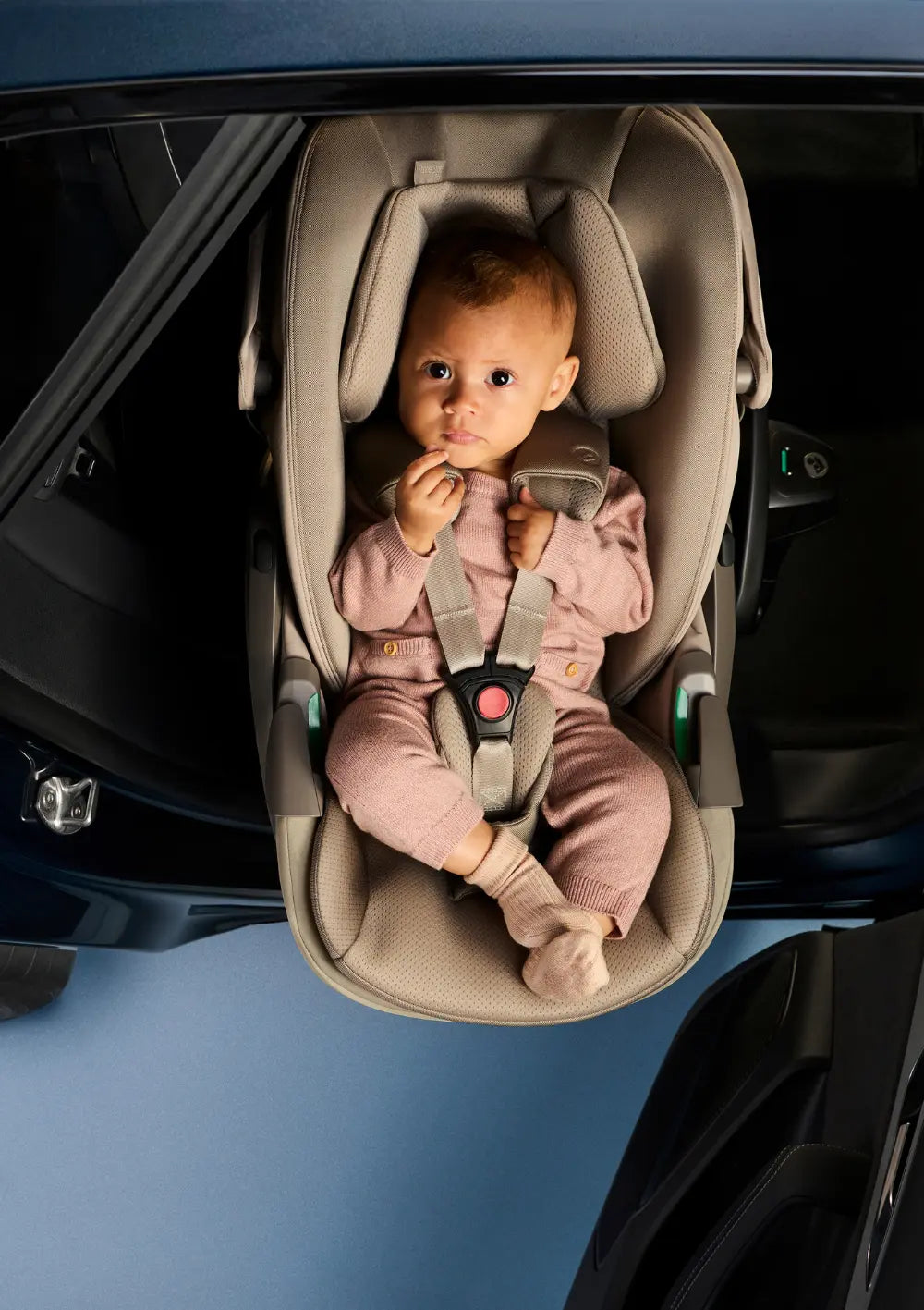 Draufsicht auf ein Baby, das in einem rosa Strampler und Söckchen in der Maxi-Cosi Pebble Slide Pro Babyschale liegt und direkt in die Kamera blickt; der 3-Punkt-Gurt ist korrekt geschlossen und die seitlichen Gurtführungen sind sichtbar.