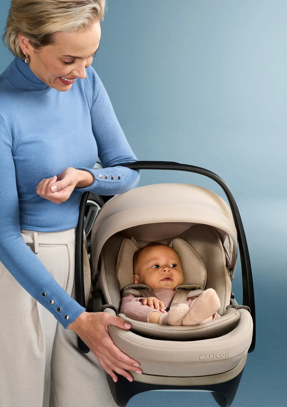 Eine lächelnde Mutter in einem blauen Rollkragenpullover blickt liebevoll auf ihr Baby, das sicher angeschnallt in der Maxi-Cosi Pebble Slide Pro Babyschale liegt, aufgenommen vor einem passenden hellblauen Studiohintergrund.