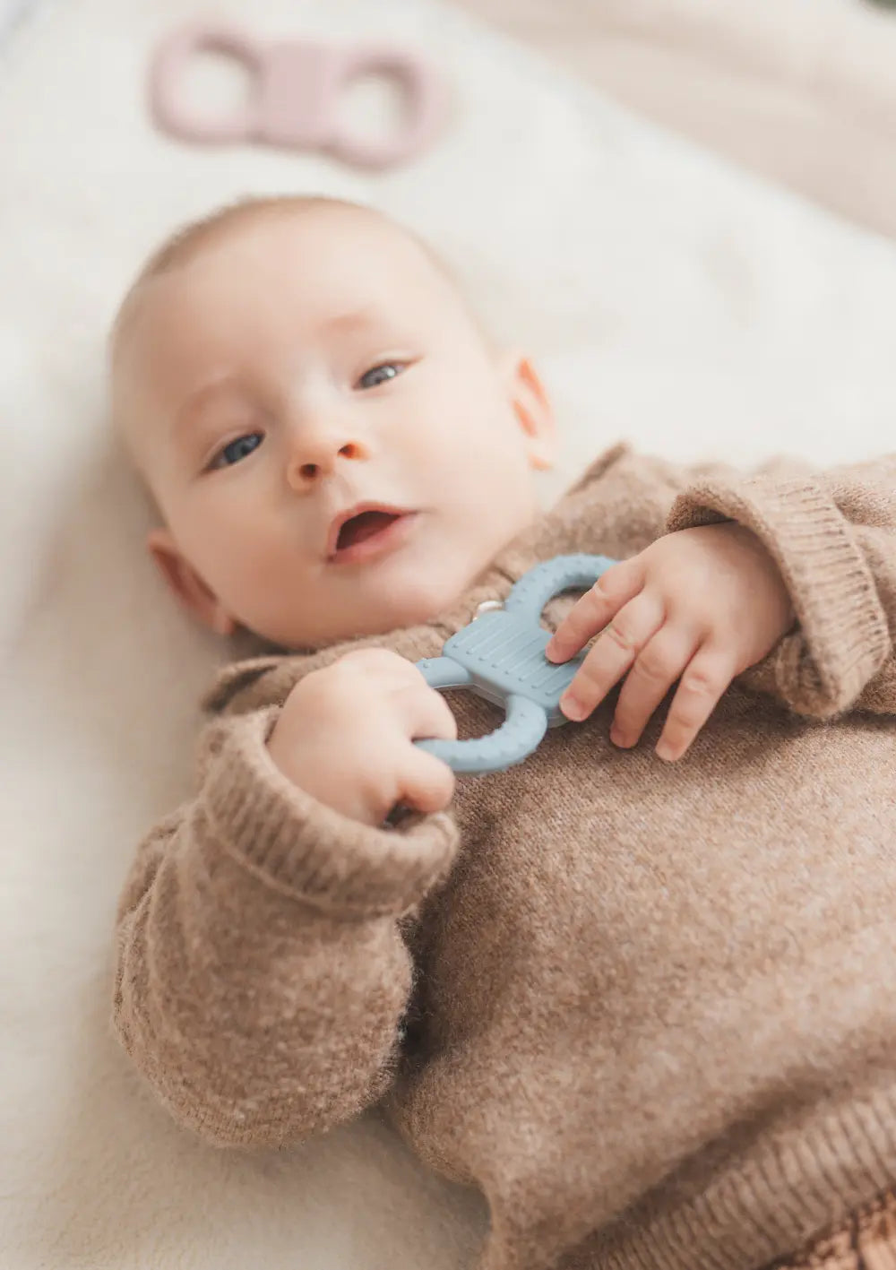 Baby liegt auf einer hellen Decke und hält den mimiloo Silikon Beißring Breeze in Blau in den Händen.