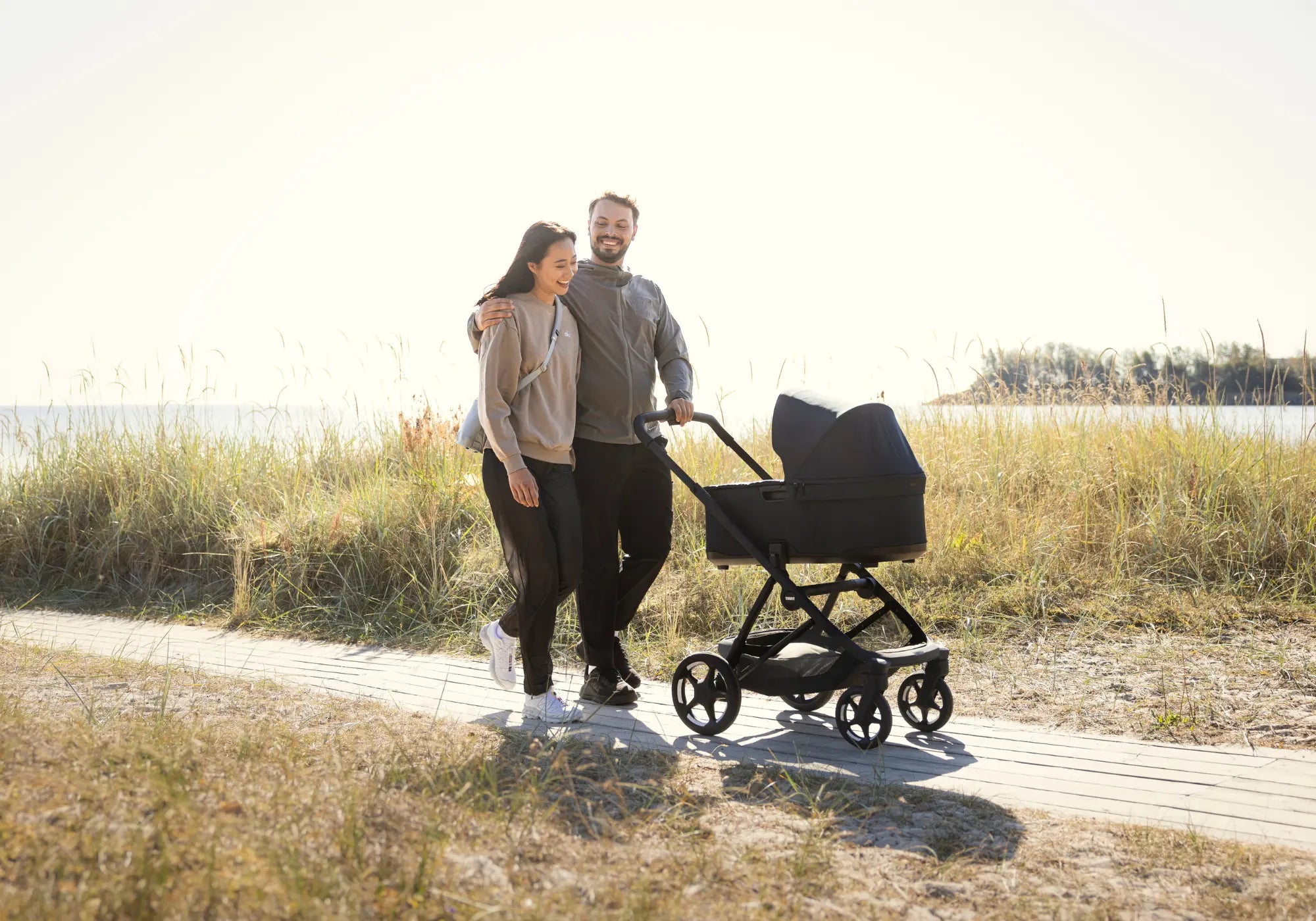 Ein lächelndes Paar spaziert auf einem Holzsteg am Meer entlang und schiebt das Kinderwagen-Set 'Charm' in Faded Khaki mit Babywanne.