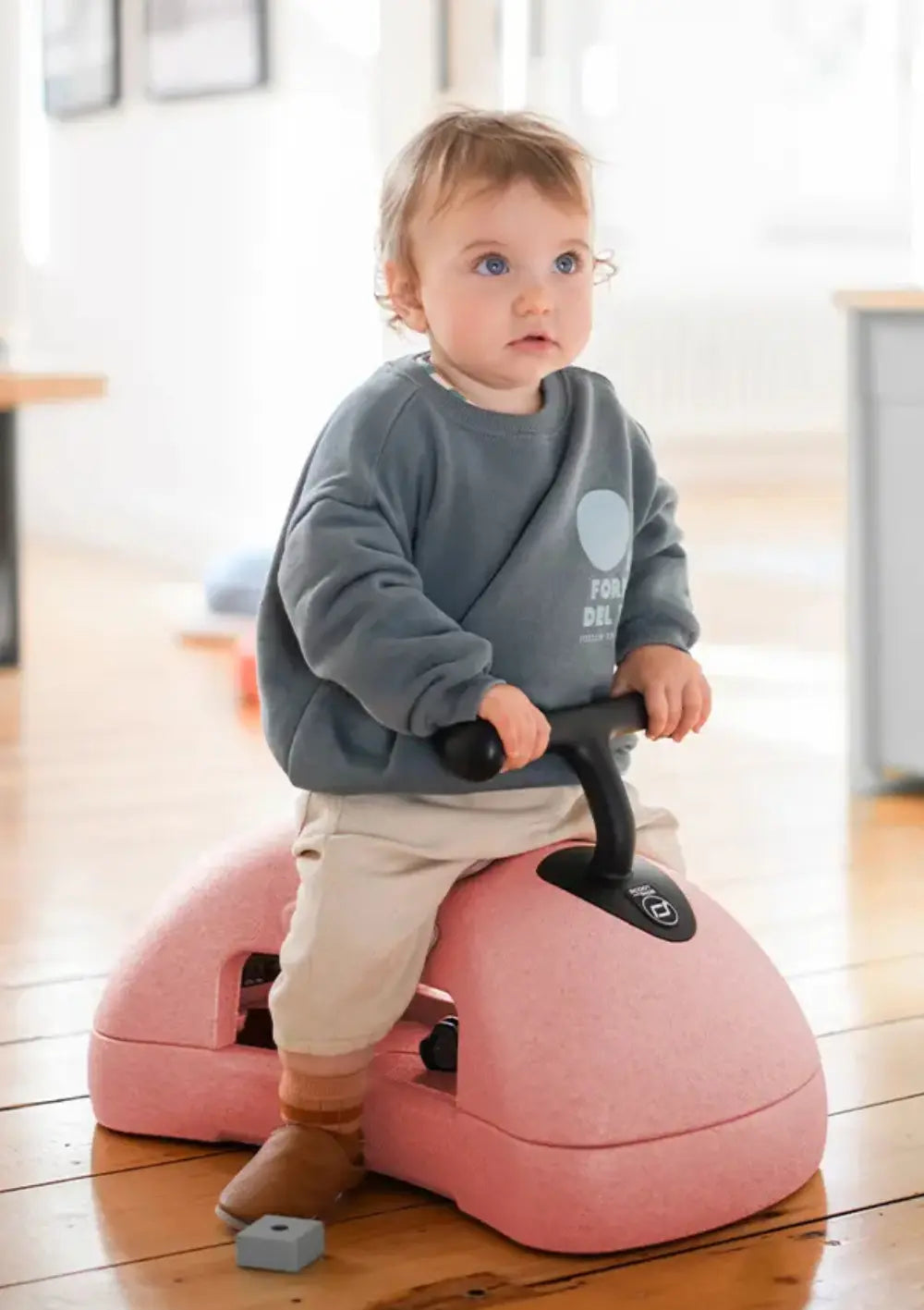 Ein Kleinkind im grauen Sweatshirt sitzt auf dem rosafarbenen Scoot and Ride 3in1 Babyroller My First Rose und hält sich sicher an den ergonomischen, schwarzen Griffen fest.