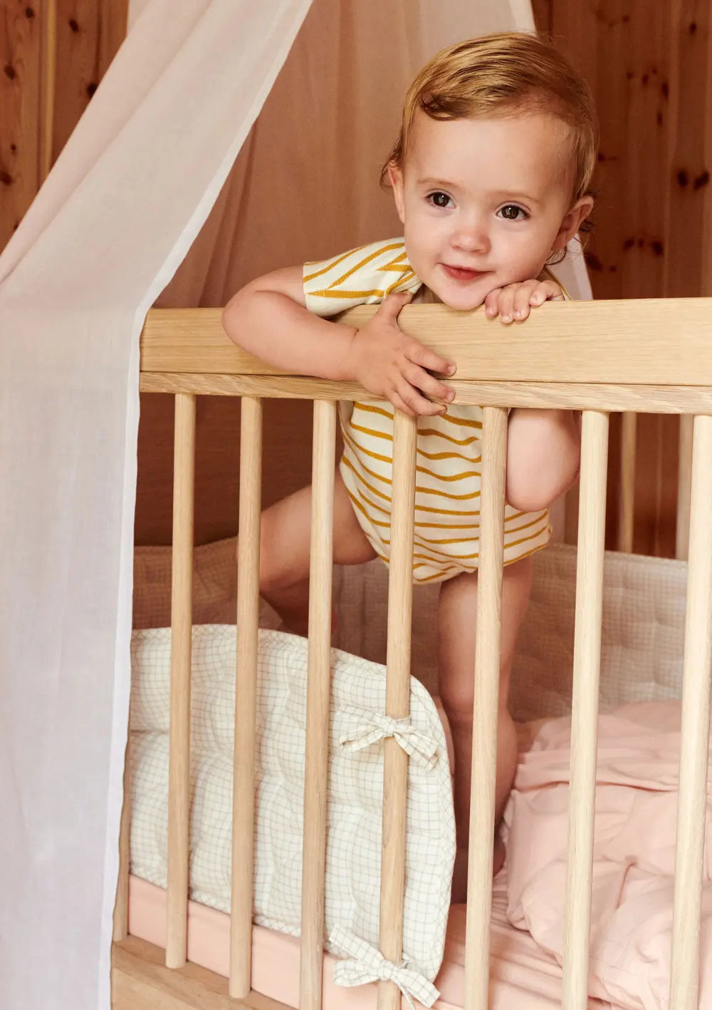 Ein Kleinkind im gestreiften Body blickt fröhlich über den Rand eines Holz-Gitterbettes, das mit dem karierten Dear April Bettnestchen Nori Mini Check ausgepolstert ist.