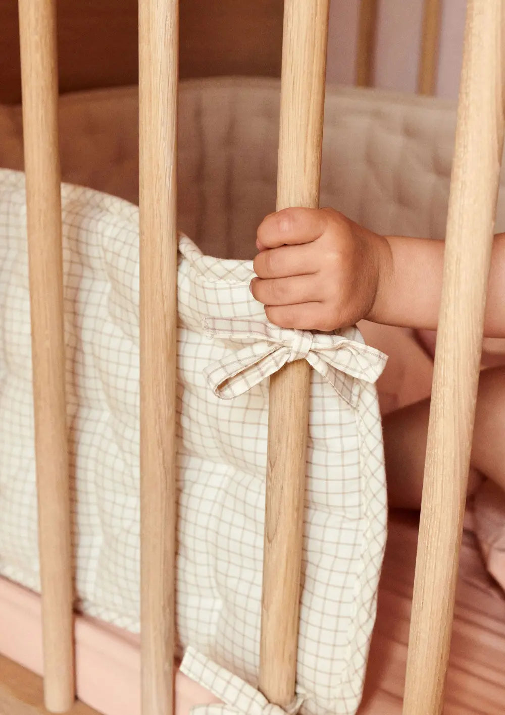 Nahaufnahme der Befestigung: Eine kleine Kinderhand greift nach den Gitterstäben des Babybettes, an denen das Dear April Bettnestchen Nori Mini Check mit einer Schleife festgebunden ist.