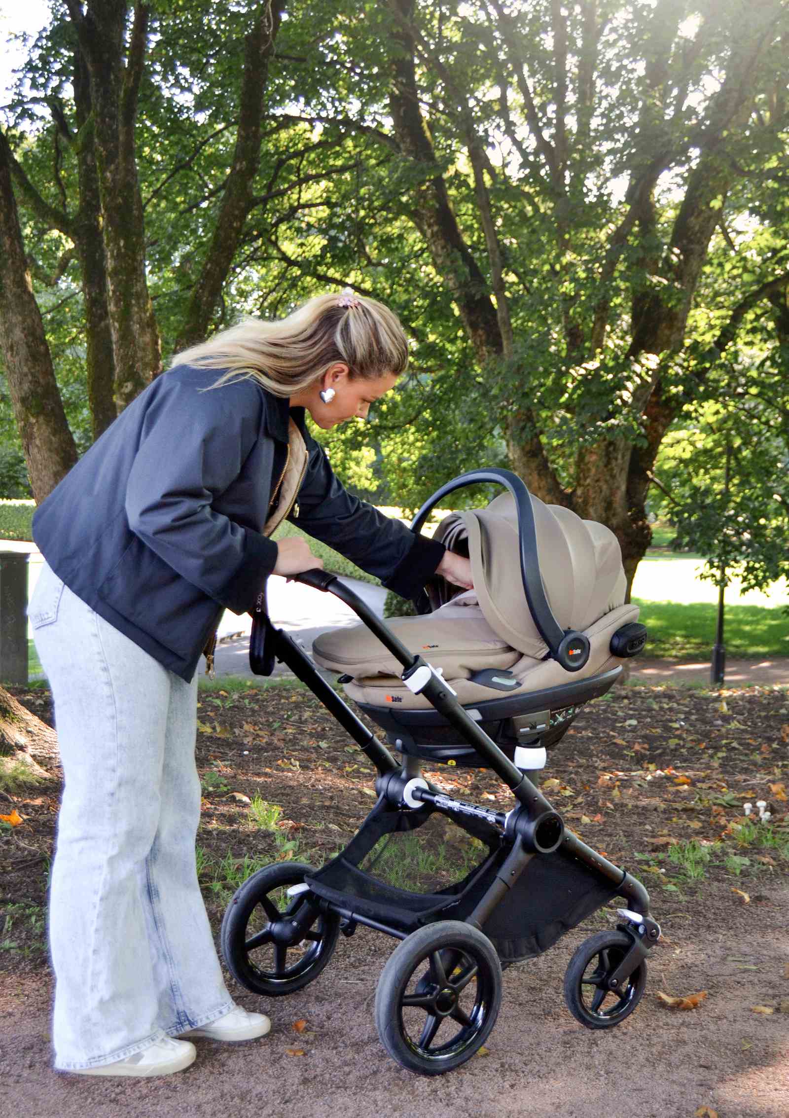 Frau schiebt am Kinderwagen befestigte BeSafe  Babyschale Go Beyond, ausgestattet mit Wind & Weather Cover Dark Sand