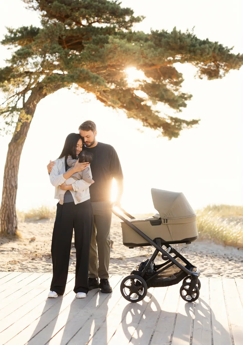 Eltern mit Baby im Arm auf einem Holzsteg am Meer; im Vordergrund steht ein Thule Kinderwagen-Set 'Charm' Faded Khaki im Gegenlicht.