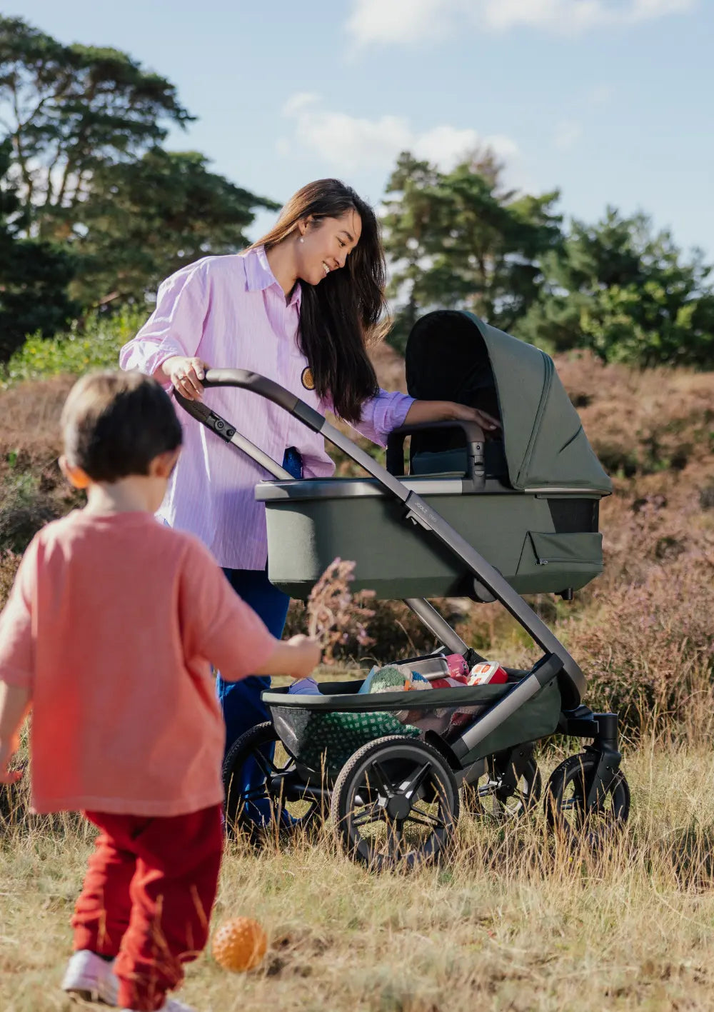 Frau schaut zu Baby in Babywanne des Joolz Kinderwagen-Sets 'Geo5' Single Forest Green – im Vordergrund: älteres Geschwisterkind