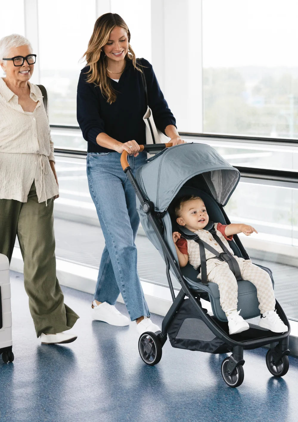 Unterwegs auf Reisen: Eine junge Frau und eine ältere Dame schieben ein Kleinkind im blaugrünen UPPAbaby Buggy Minu V3 Dillan durch eine moderne, helle Wartehalle.