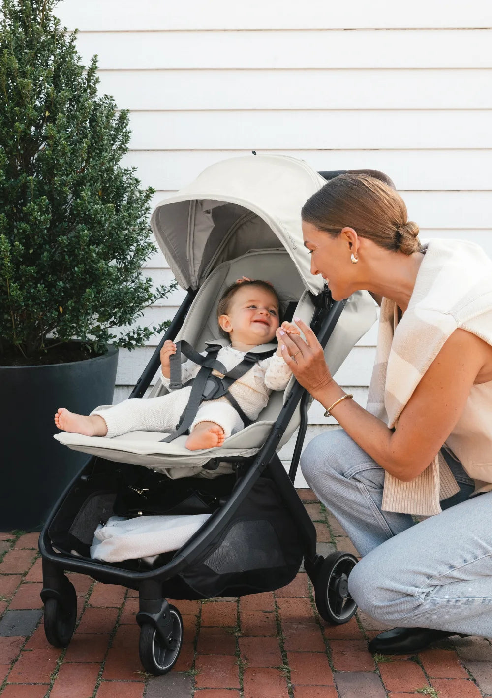 Eine Mutter kniet lächelnd vor dem cremefarbenen UPPAbaby Buggy Minu V3 Savannah und hält die Hand ihres Babys; helle Hauswand im Hintergrund.