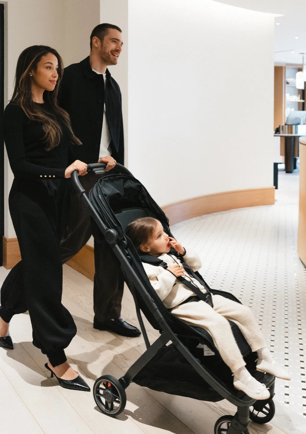 Ein Paar spaziert durch einen eleganten Innenraum und schiebt ein Kleinkind im schwarzen UPPAbaby Buggy Minu V3 James.
