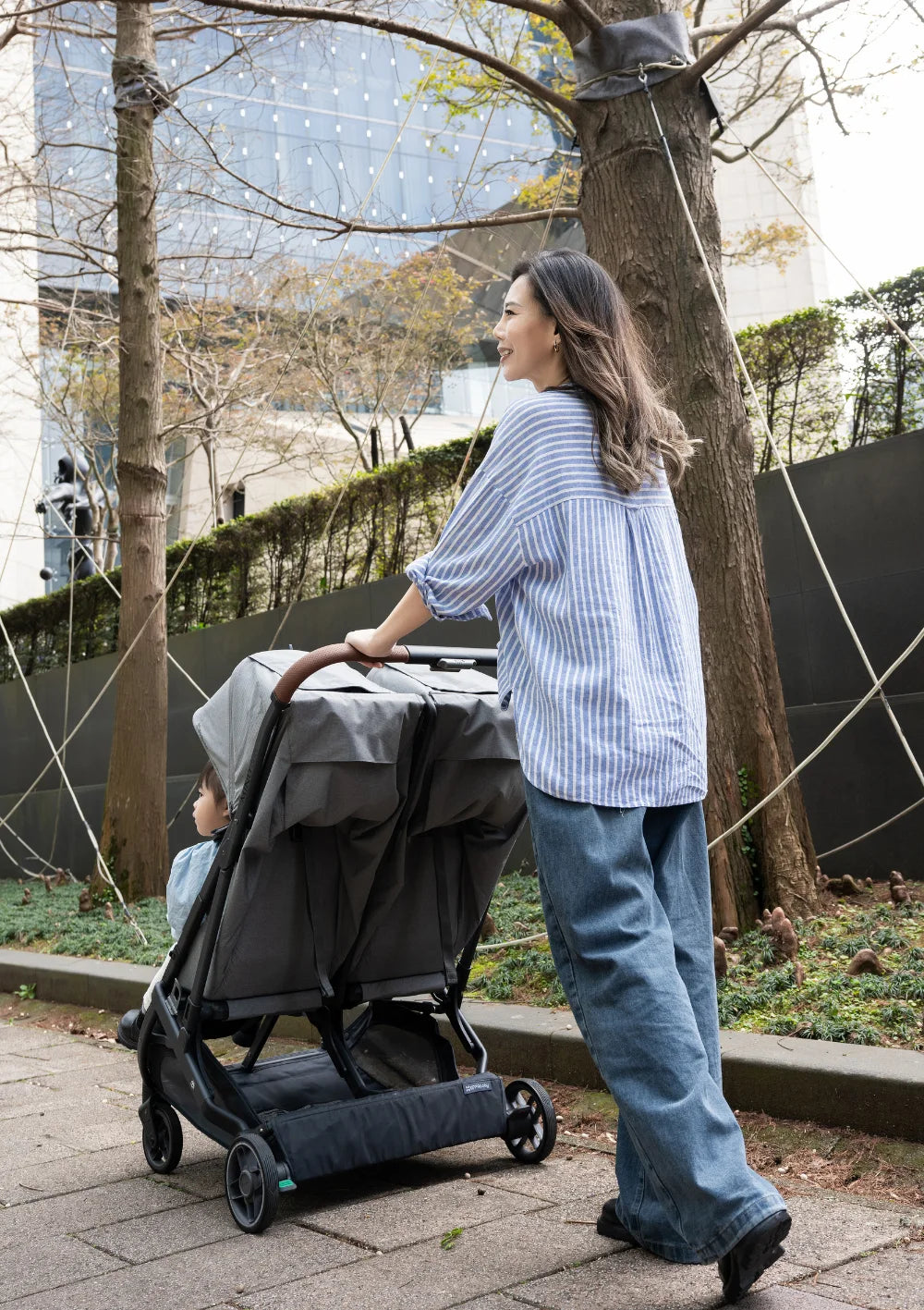 Eine Frau schiebt den grauen UPPAbaby Geschwisterbuggy Minu Duo Greyson auf einem gepflasterten Weg an Bäumen und einer modernen Glasfassade vorbei.