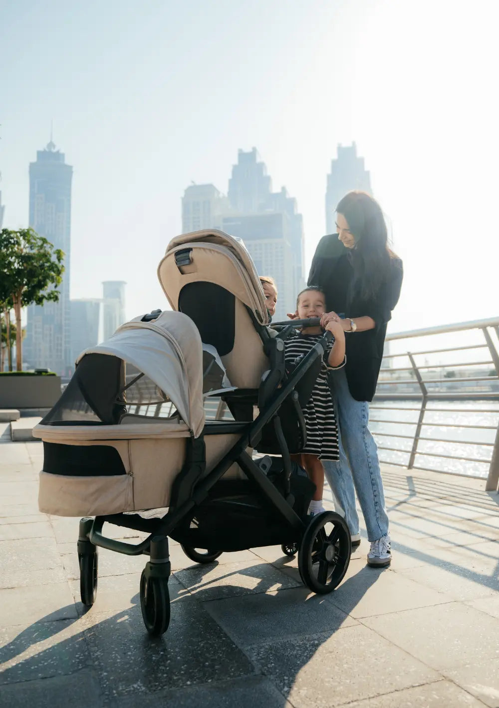 Eine Frau ist mit drei Kindern an einer Uferpromenade in der Stadt unterwegs. Im Bild: das Kinderwagen-Set UPPAbaby 'Vista 3' Liam mit Babywanne,   Sportsitz und Mitfahrbrett