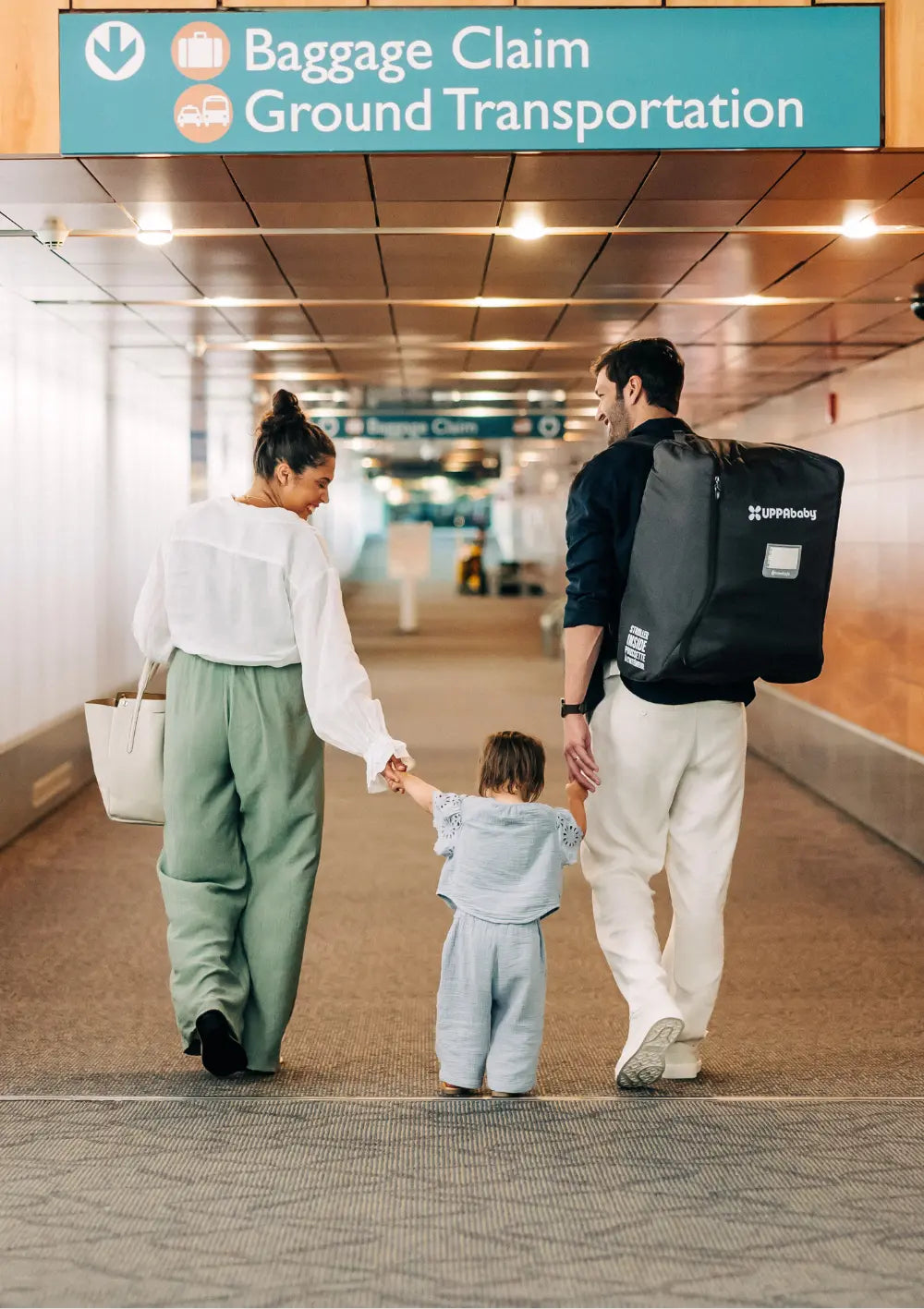 Rückansicht einer Familie mit Kleinkind am Flughafen unter einem Schild für Gepäckausgabe. Der Mann trägt die UPPAbaby Reisetasche für Minu V3 mit Rucksackgurten, um die Hände frei zu haben.