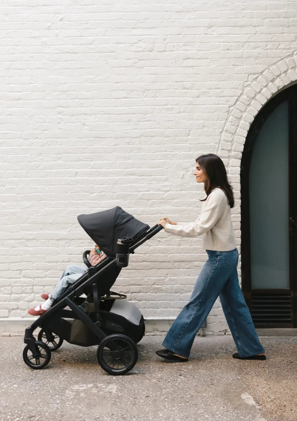 Seitenansicht einer Frau beim Spaziergang mit einem schwarzen UPPAbaby Kinderwagen. Am Gestell ist der praktische UPPAbaby Becherhalter befestigt, in dem ein Thermobecher steckt.