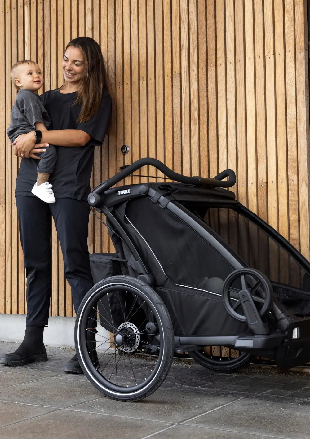Eine lächelnde Mutter hält ihr Kind neben dem Thule Multisport-Fahrradanhänger Chariot Sport 2 Single Black vor einer modernen Holzfassade.