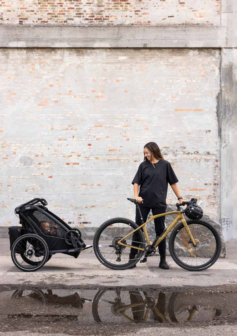 Der Thule Multisport-Fahrradanhänger Chariot Sport 2 Single Black als Fahrradanhänger an einem Fahrrad vor einer rustikalen Backsteinwand.