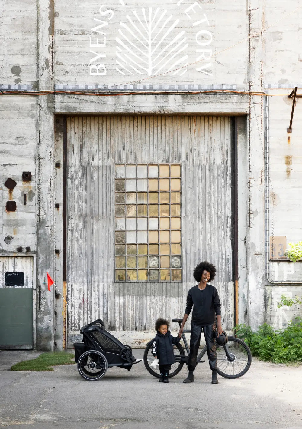 Mutter und Kind stehen mit dem Thule Multisport-Fahrradanhänger Chariot Sport 2 Single Black vor einem großen Industriegebäude.