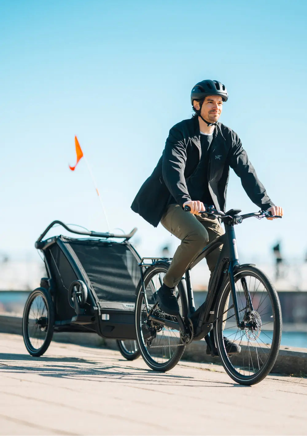 Ein Mann fährt ein E-Bike mit dem angekoppelten Thule Multisport-Fahrradanhänger Chariot Sport 2 Double Black auf einem sonnigen Weg am Wasser entlang; sportliche Freizeitgestaltung.