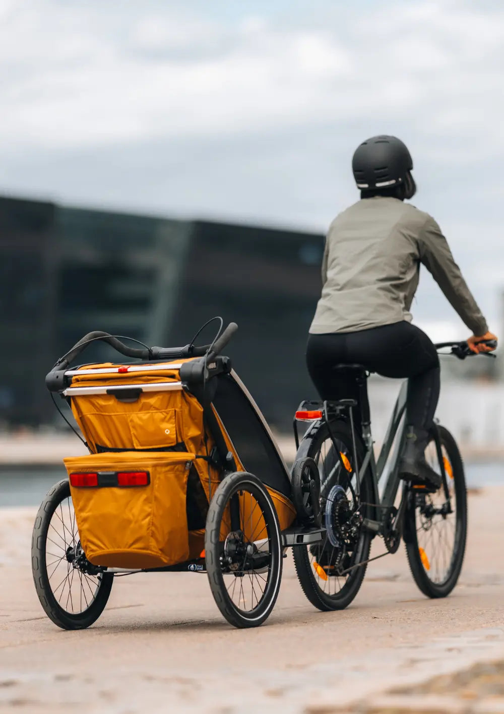 Rückansicht des Thule Multisport-Fahrradanhängers Chariot Sport 2 Single Natural Gold im Einsatz als Fahrradanhänger; Fokus auf die goldene Farbe und die Sicherheitsreflektoren.