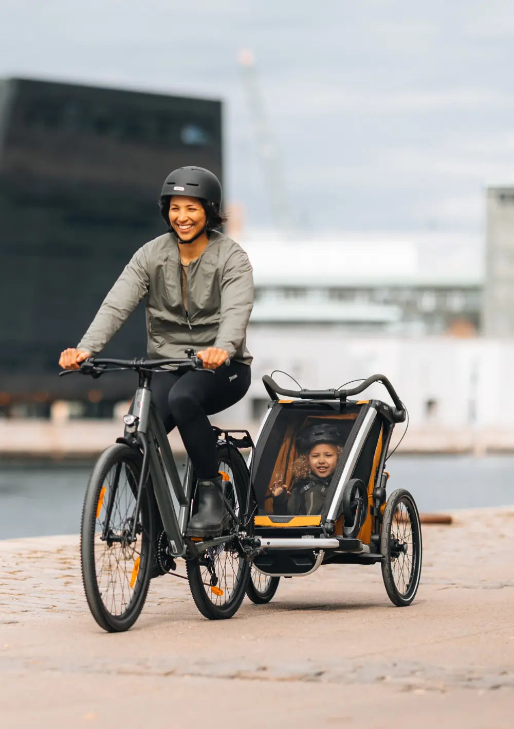 Eine lachende Frau fährt mit dem Thule Multisport-Fahrradanhänger Chariot Sport 2 Single Natural Gold entlang einer modernen Uferpromenade; urbaner Multisport-Einsatz.