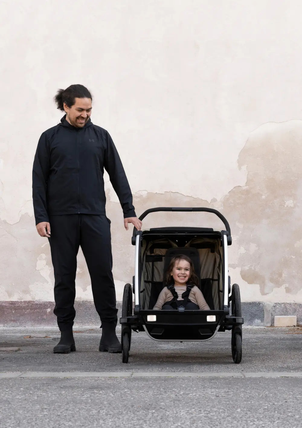 Ein lächelndes Kind sitzt im Thule Multisport-Fahrradanhänger Chariot Cross 2 Double Faded Khaki, während der Vater den Anhänger im Buggy-Modus hält; moderne Outdoor-Aufnahme.