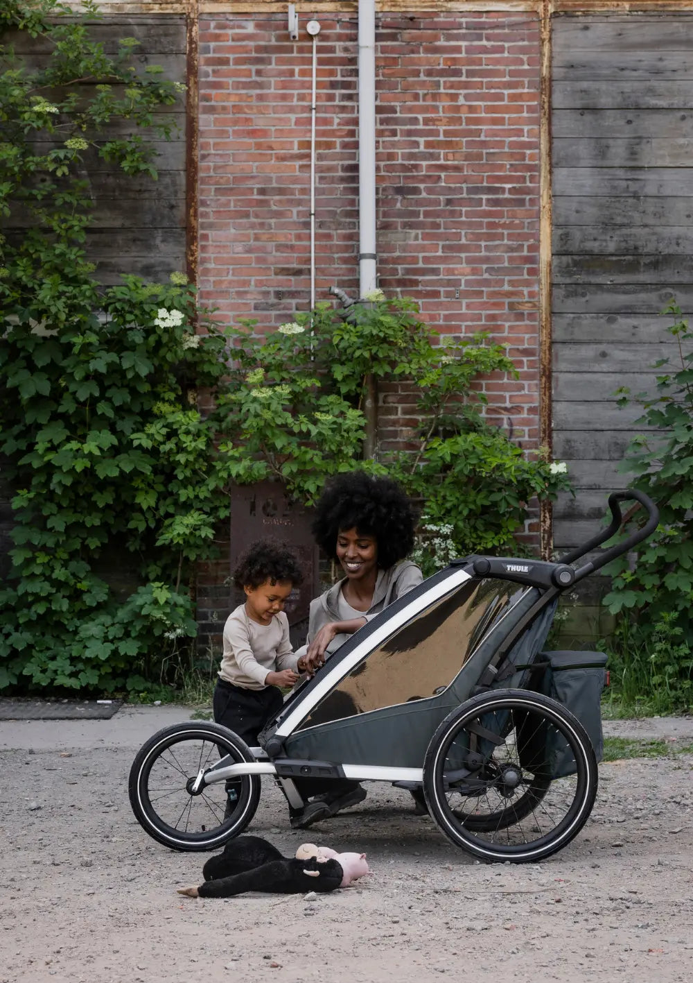 Seitenprofil des Thule Chariot 2 auf Schotterboden; eine Mutter und ihr Kind stehen am Wagen, an dem das Thule All-Terrain Kit für Fahrradanhänger Chariot 2 Double für Outdoor-Aktivitäten angebracht ist.