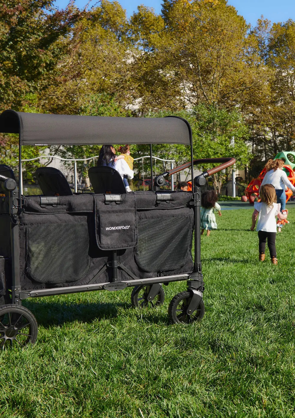 Der WonderFold W6 Luxe Pro Kinderwagen 6-Sitzer Jet Schwarz mit schützendem Sonnendach steht auf einem Rasen neben einem Spielplatz.