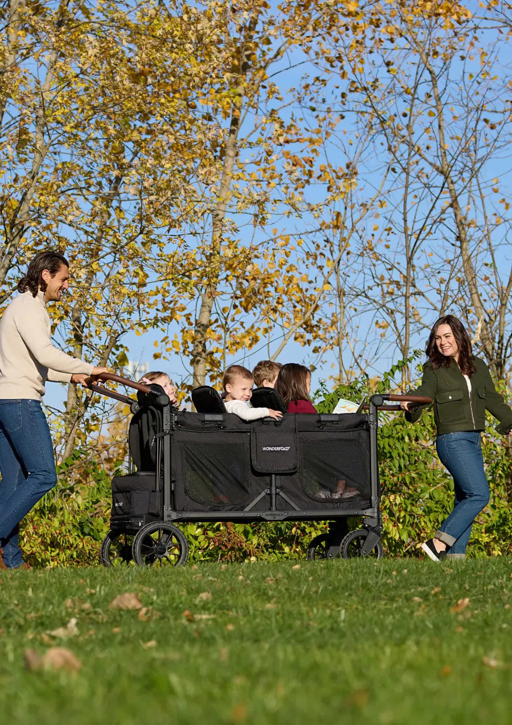 Ein Paar schiebt gemeinsam den WonderFold W6 Luxe Pro Kinderwagen 6-Sitzer Jet Schwarz über eine Wiese; Platz für bis zu sechs Kinder bei Familienausflügen in der Natur.
