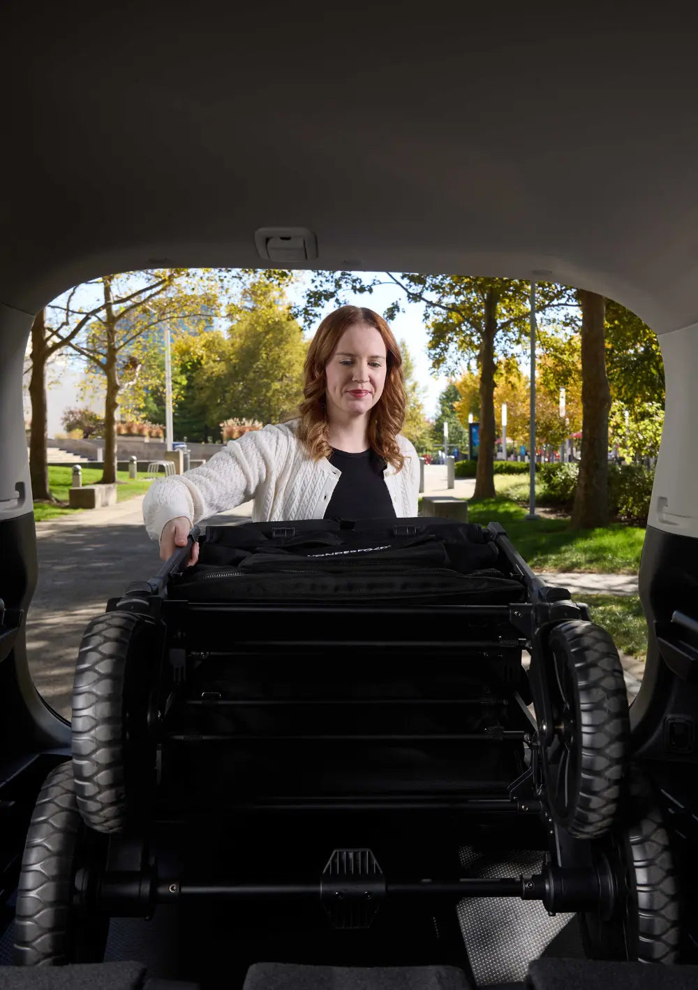 Eine Frau verstaut den zusammengeklappten WonderFold W4 Luxe Pro Kinderwagen 4-Sitzer Jet Schwarz mühelos im Kofferraum eines Autos.