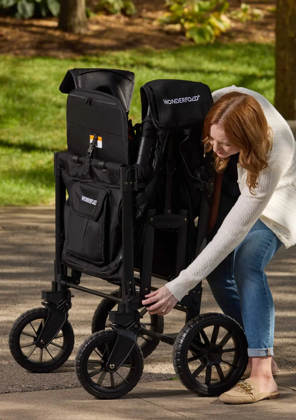 Eine Frau zeigt, wie kompakt sich der WonderFold W4 Luxe Pro Kinderwagen 4-Sitzer Jet Schwarz für den Transport oder die Lagerung zusammenfalten lässt.