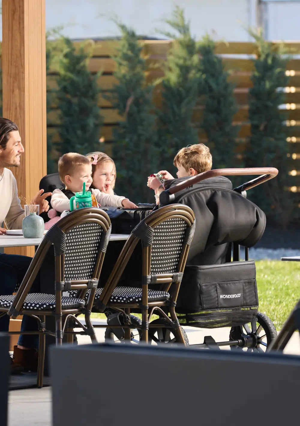 Der WonderFold W4 Luxe Pro Kinderwagen 4-Sitzer Jet Schwarz dient als praktischer Hochstuhlersatz am Esstisch im Freien, während drei Kinder darin sitzen.