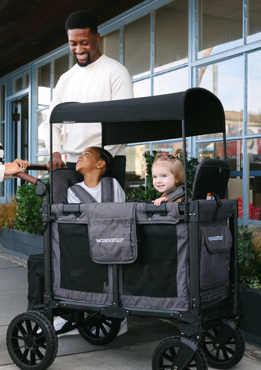 Ein Vater schiebt zwei Kinder im WonderFold W2 Luxe Pro Kinderwagen 2-Sitzer Jet Schwarz auf einem Gehweg vor einer Fensterfront; urbaner Familienausflug.