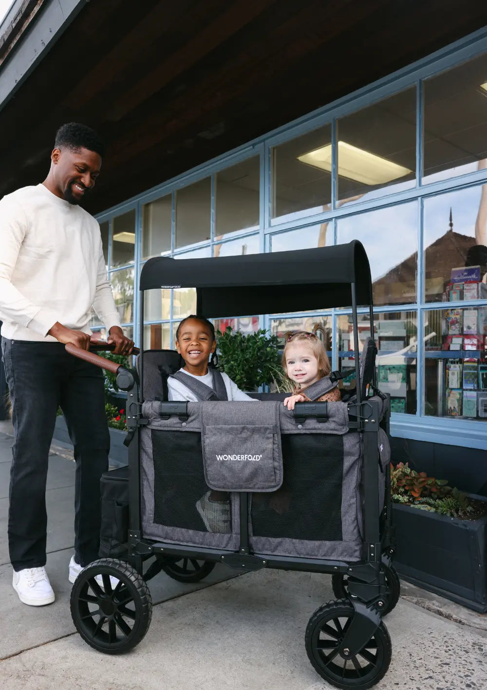 Ein Vater schiebt zwei Kinder im WonderFold W2 Luxe Pro Kinderwagen 2-Sitzer auf einem Gehweg vor einer Fensterfront; urbaner Familienausflug.