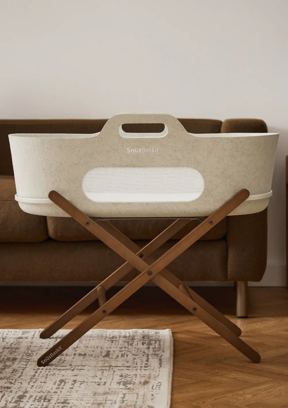 Der hellbeige Snüz Moseskorb inkl. Ständer SnüzBaskit Barley-Walnuss steht komplett aufgebaut auf seinem dunkelbraunen Holz-Klappgestell im Wohnzimmer vor einem Sofa.