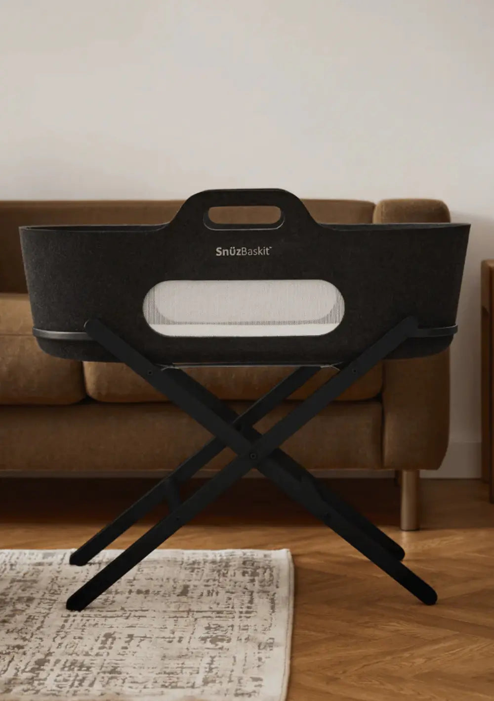 Der anthrazitfarbene Snüz Moseskorb inkl. Ständer SnüzBaskit Graphite steht standsicher auf einem schwarzen Holz-Klappgestell vor einem braunen Sofa.