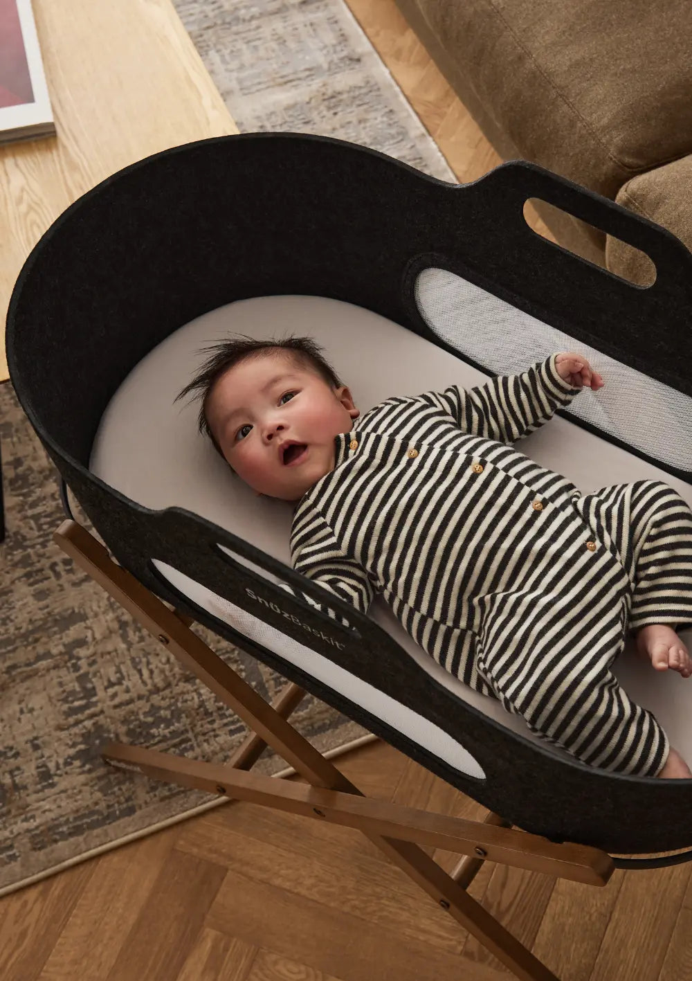 Blick von oben auf ein liegendes Baby im anthrazitfarbenen Snüz Moseskorb inkl. Ständer SnüzBaskit Graphite-Walnuss mit komfortabler, weißer Matratze.