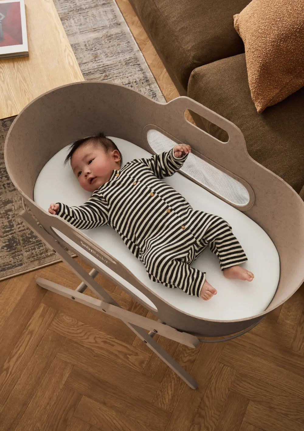 Ein Baby im gestreiften Strampler liegt gemütlich auf der weichen Matratze im Snüz Moseskorb inkl. Ständer SnüzBaskit Mocha; Draufsicht auf den sicheren Schlafplatz.