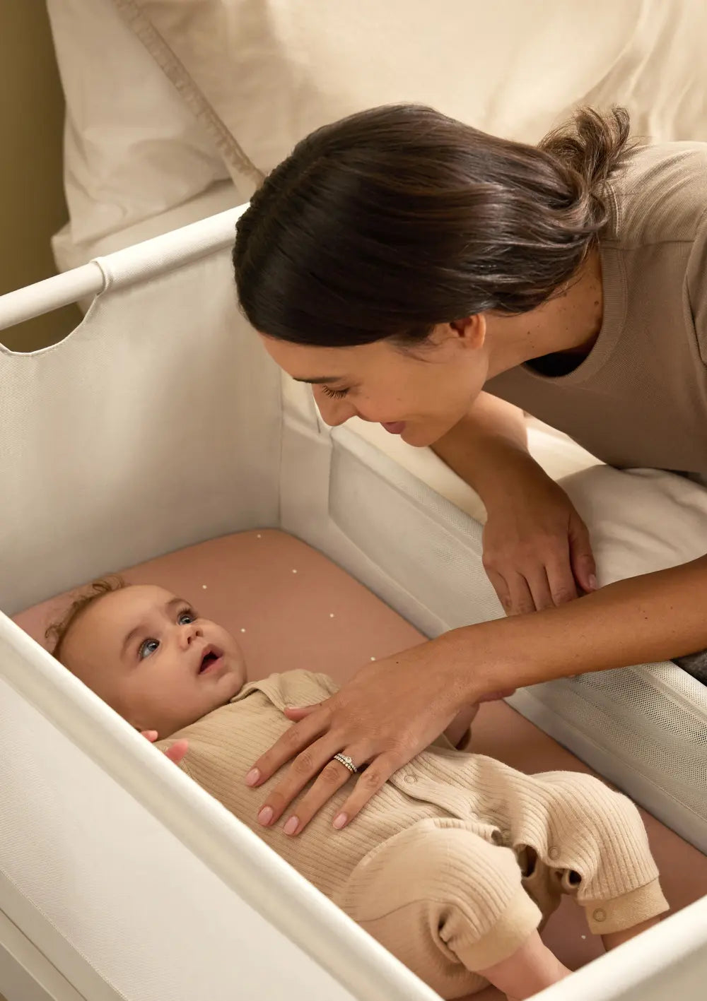Eine Mutter blickt liebevoll auf ihr im Snüz Beistellbett SnüzPod 5 Cashmere liegendes Baby und legt ihm sanft die Hand auf den Bauch.